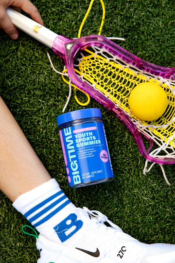 Lacrosse stick with a yellow ball on grass, next to a container of Big Time Youth Sports Gummies