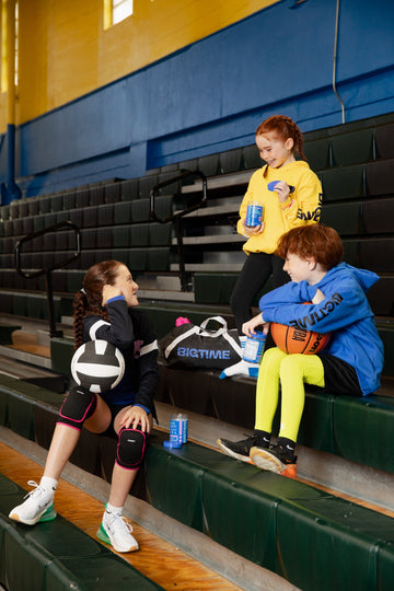Children with sports equipment sitting on bleachers in a gymnasium.